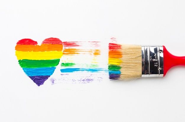 A rainbow-colored heart made of paint swipes with a paintbrush in color-striped rainbow hues to the right, some spilling a...